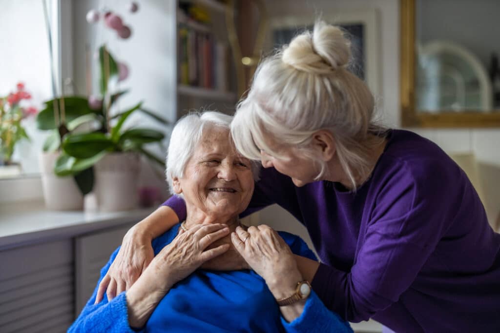 Woman hugs sitting elderly woman from behind as they smile.