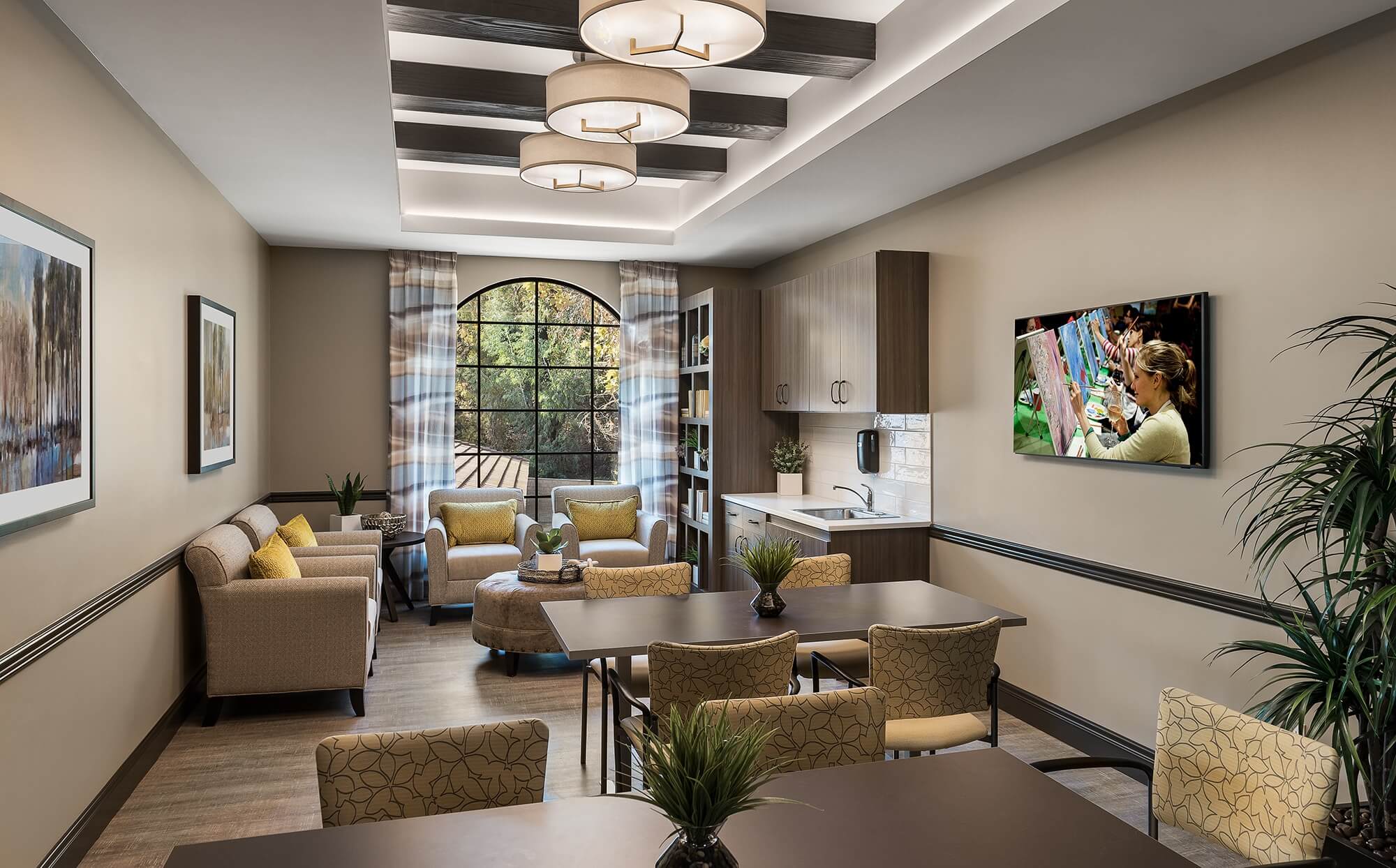 Bright activity room with tables and chairs, and comfortable seating near a kitchenette and a large window.